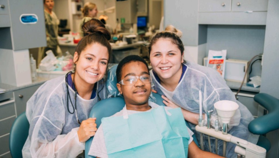 Two dental hygiene students pose with a young patient in the Harcum College Dental Clinic, all smiling during a dental visit.