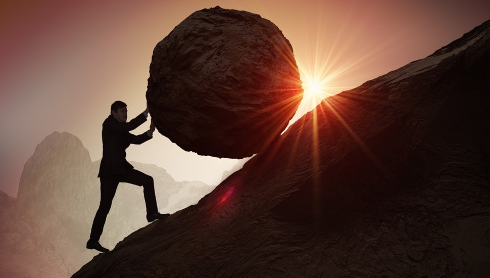 Silhouette of businessman pushing heavy stone boulder up on hill.