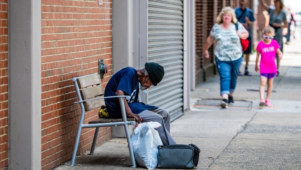 An elderly homeless man sitting on a bench by a red brick wall with people walking past in the background.