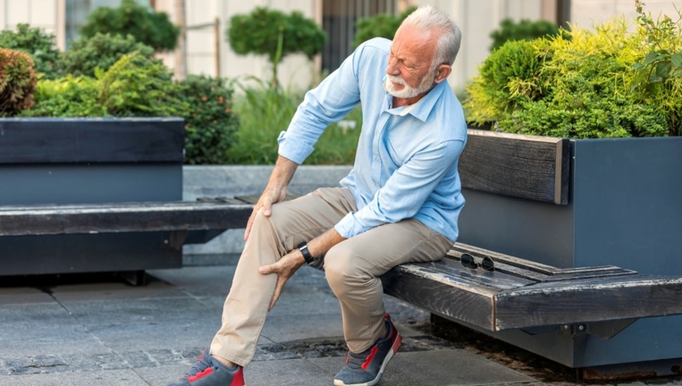 Old man sits on bench and holds his knee in pain.