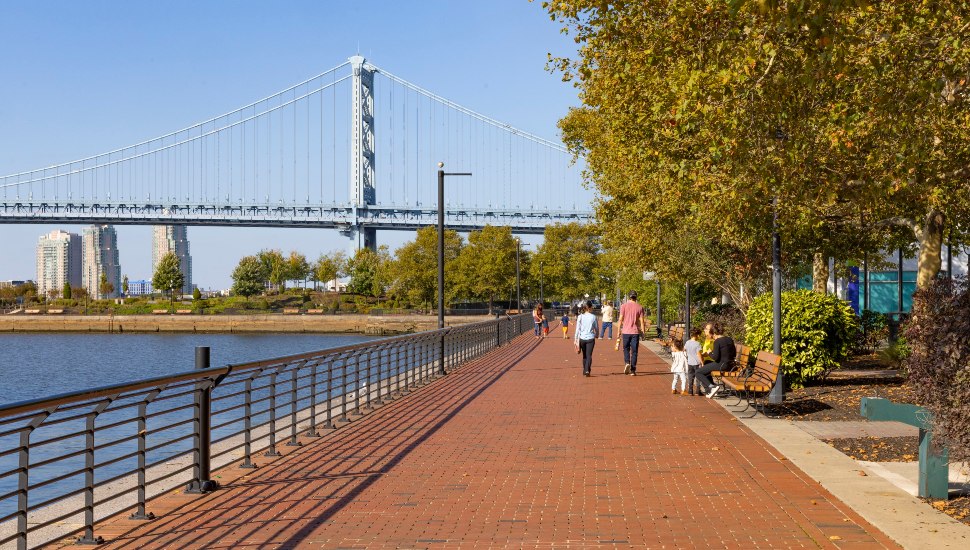 People walk on trail alongside river with bridge in background.