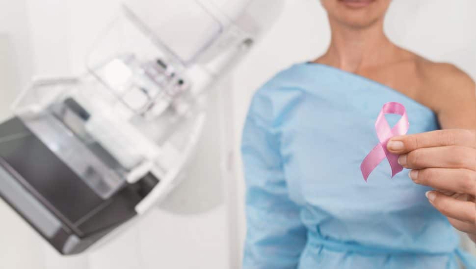 Woman holding breast cancer pink ribbon next to special medical equipment mammograph.