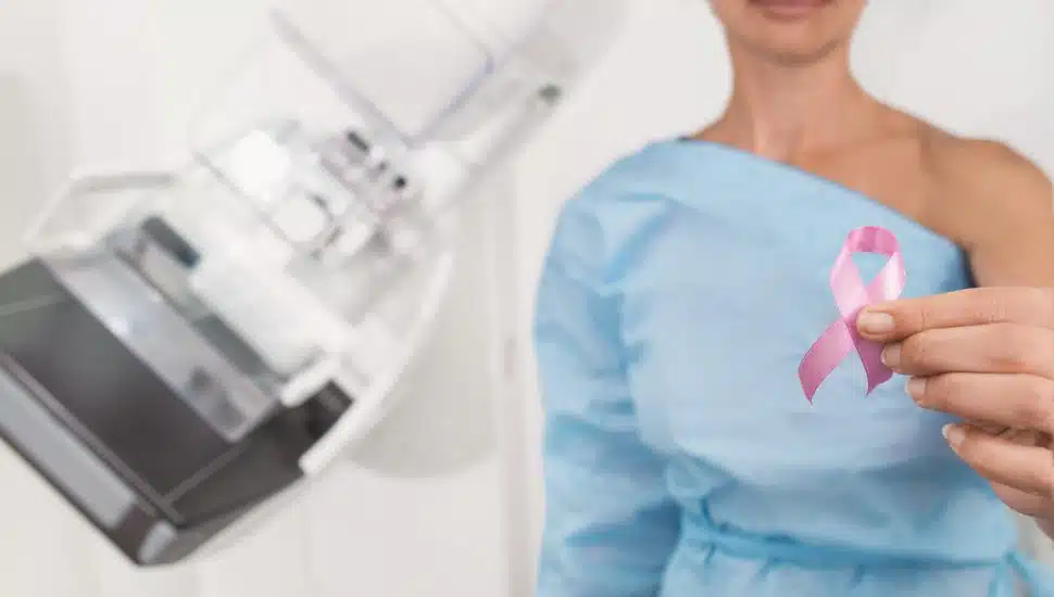 Woman holding breast cancer pink ribbon next to special medical equipment mammograph.