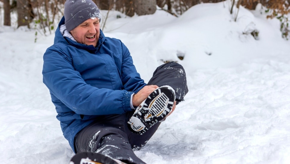 Man sits in snow and grabs ankle in pain.