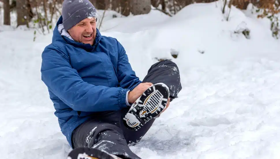 Man sits in snow and grabs ankle in pain.