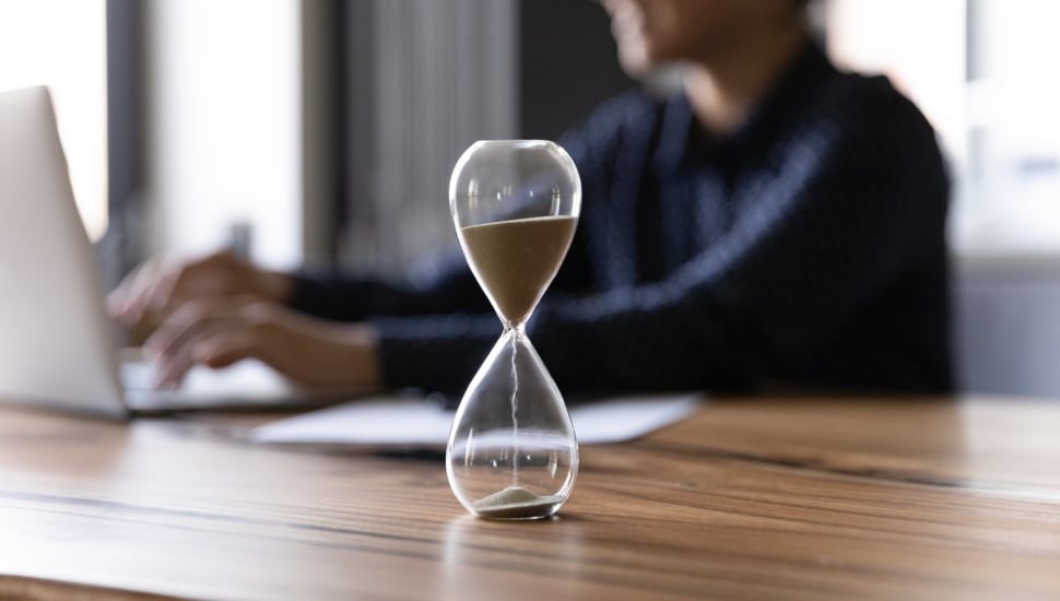 Hourglass measures time as businesswoman works in background.