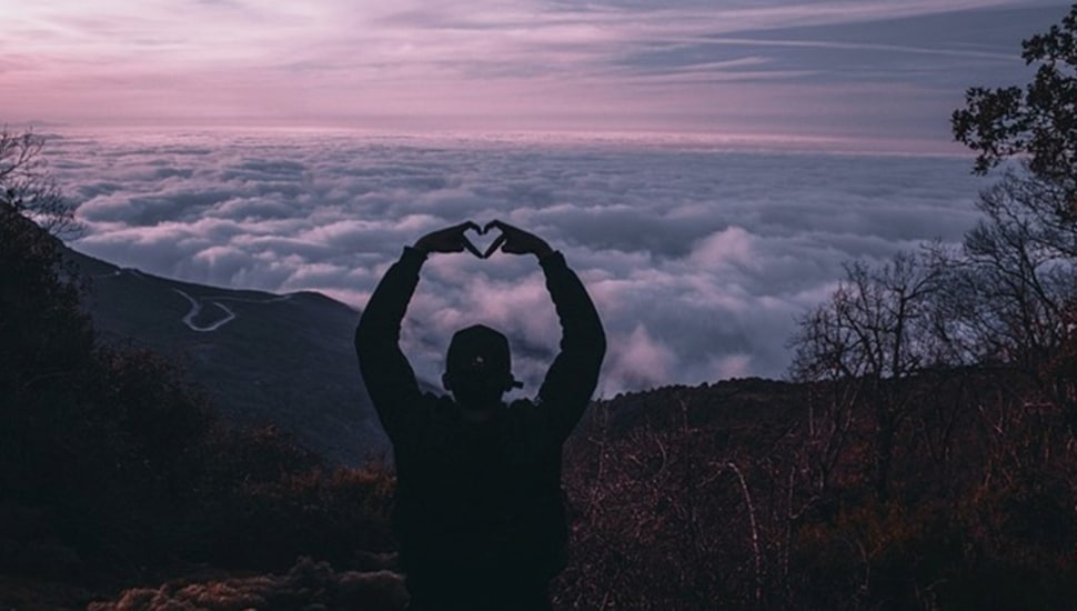 Person atop mountain makes a heart symbol with hands.