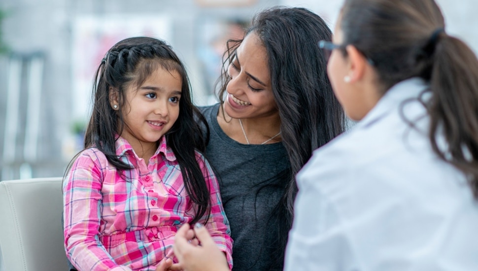 Young girl sits on mother's lap and talks to doctor.