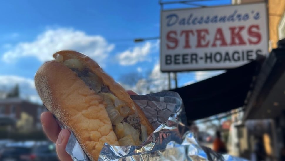 A cheesesteak being held up, half-covered in aluminum foil, outside of Dalessandro's Steaks.
