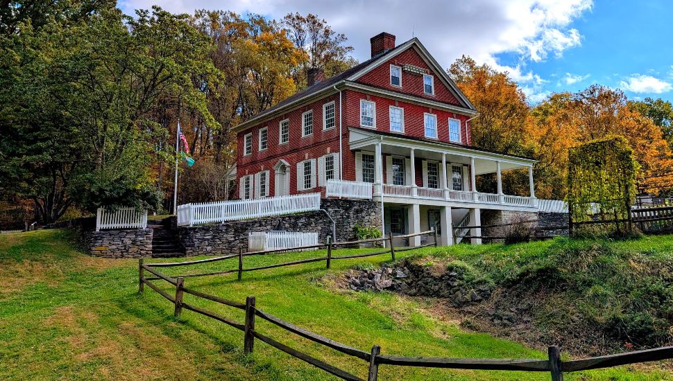 Historic Rock Ford exterior view surrounded by autumn trees, featuring the historic red brick mansion, wraparound porch, stone retaining walls, and sloping grassy landscape.