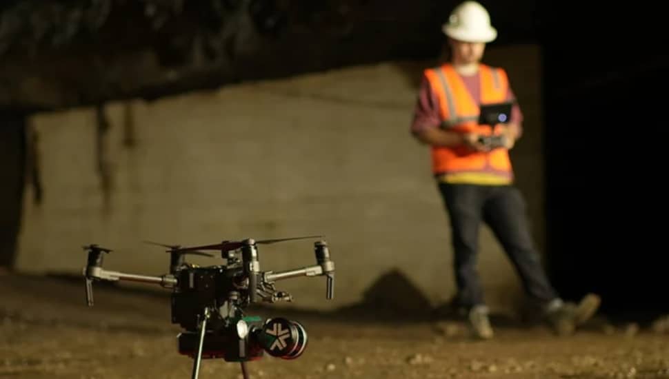 An Exyn Technologies autonomous drone in the foreground inside a dark underground environment, with a worker in an orange safety vest and hard hat holding a controller in the background.