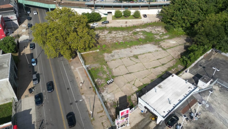 Aerial view of a vacant, cracked-concrete lot on Chelten Avenue in Germantown, Philadelphia, with the elevated Germantown Station rail structure visible in the background and a gas station at the corner.